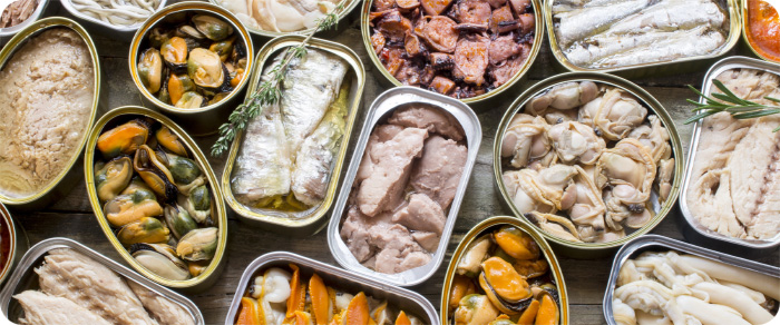 Vista cenital de un surtido de latas de conservas gourmet con mejillones, sardinas, atún y 
                    almejas sobre una superficie de madera rústica.